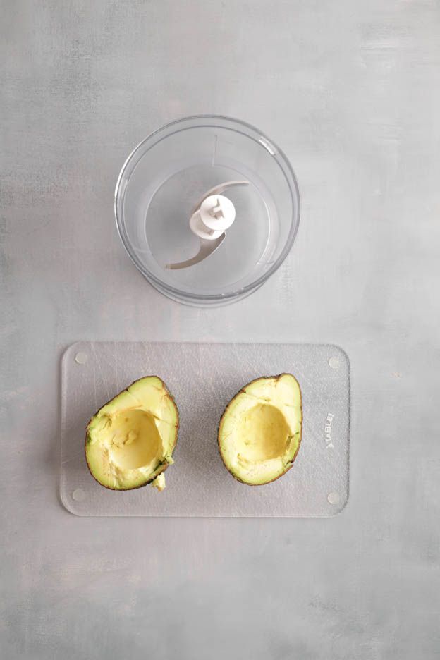 Scooped out avocado halves on a glass board next to the empty blender bowl