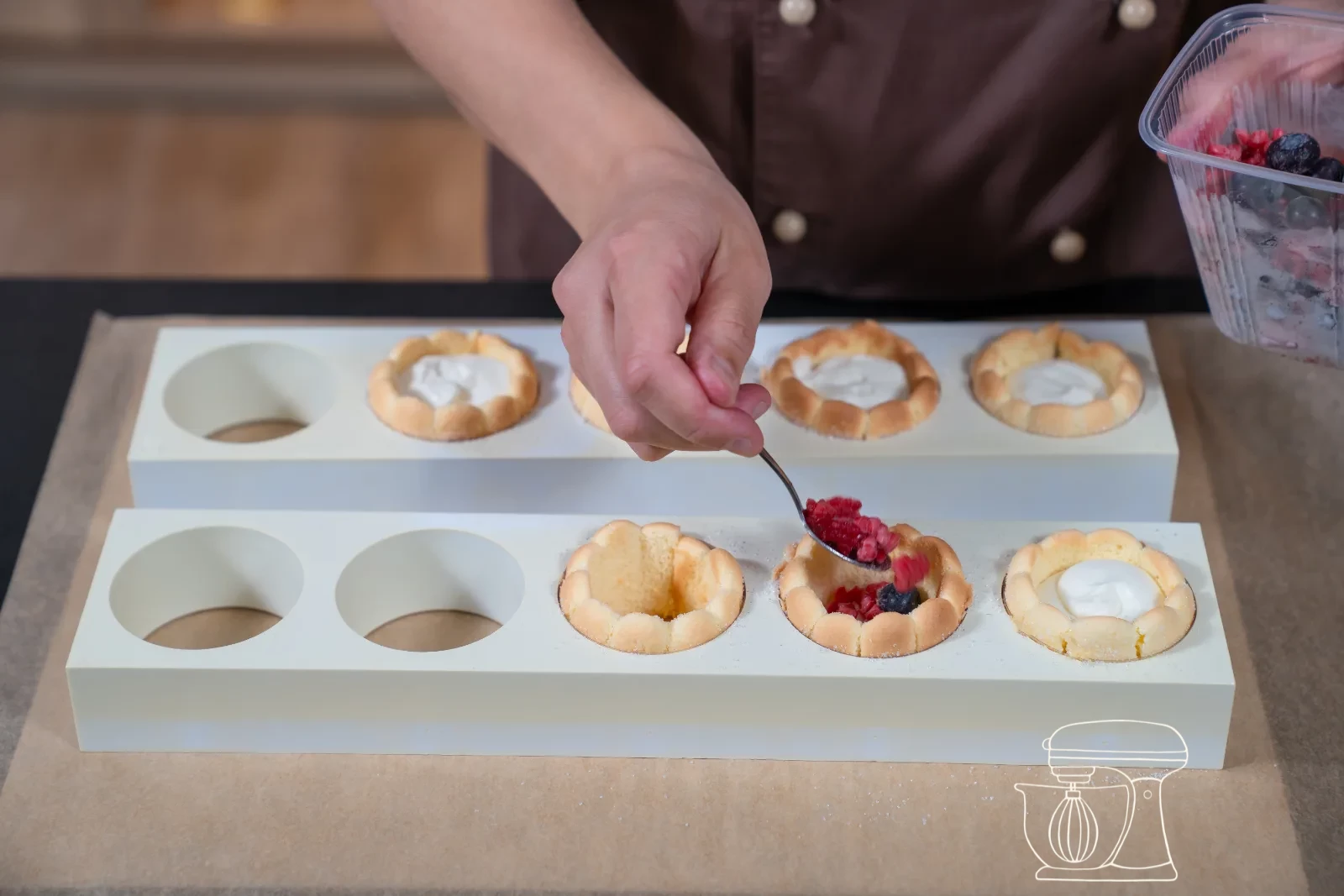 Vanilla cream being piped as a rosette onto a filled mini charlotte, with fresh berries and icing sugar in the background