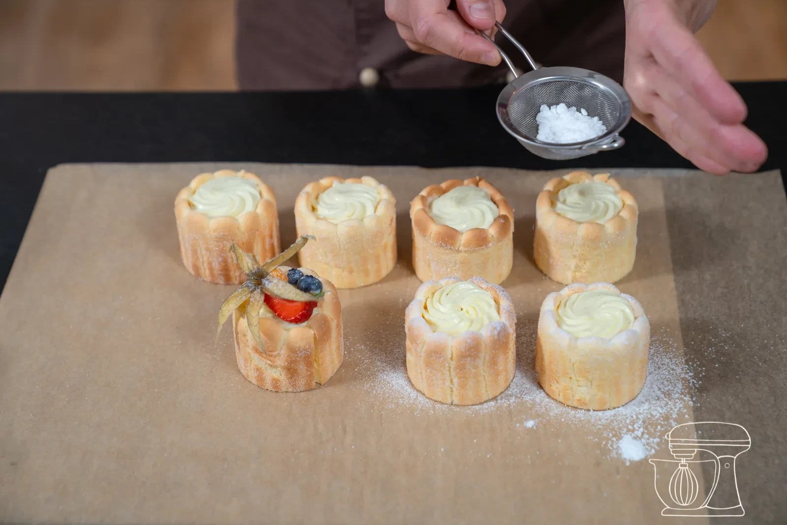 Vanilla cream being piped as a rosette onto a filled mini charlotte, with fresh berries and icing sugar in the background
