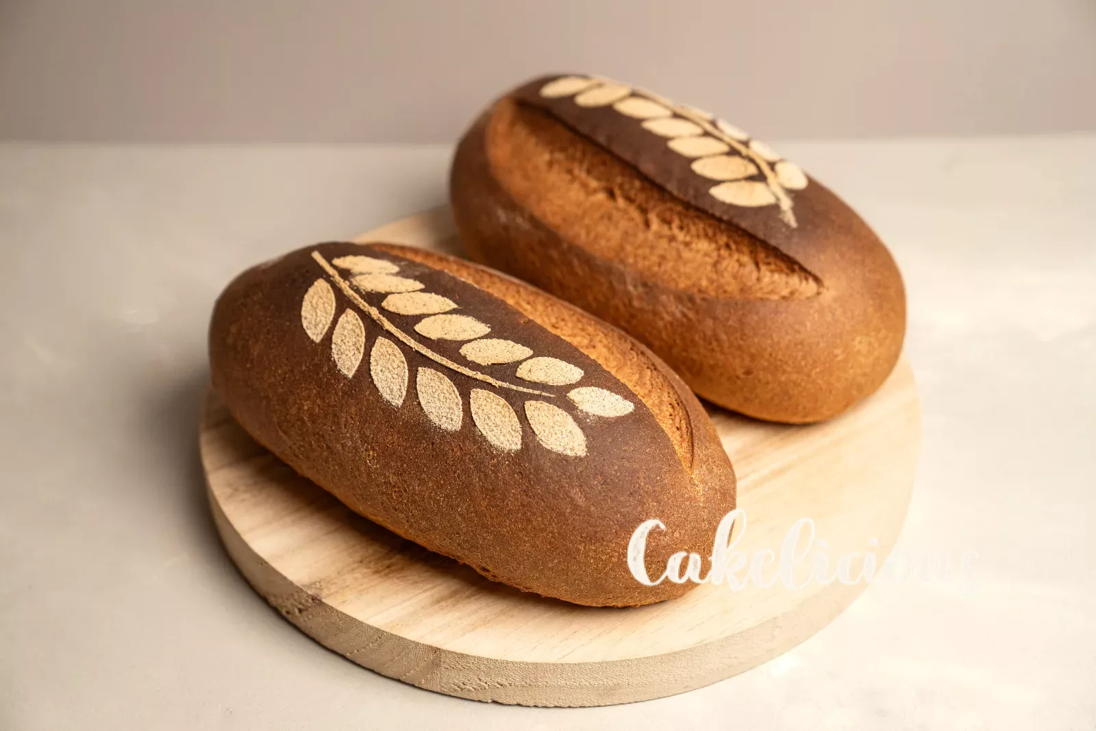 Pane rustico con motivo a spiga di farina, davanti fette tagliate su un tagliere di legno rotondo