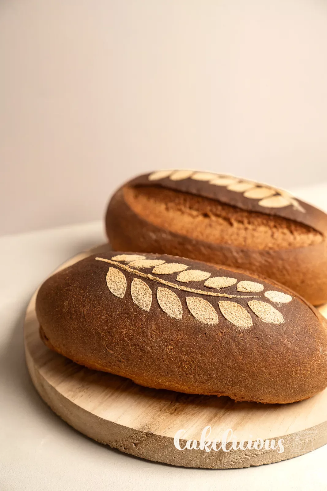 Pane rustico con motivo a spiga di farina, davanti fette tagliate su un tagliere di legno rotondo